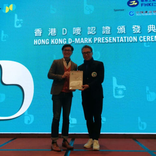 Two men standing on a stage at the Hong Kong D-Mark Presentation Ceremony. One man is handing a certificate to the other. The background features a large screen with the event's name and logos of sponsors.