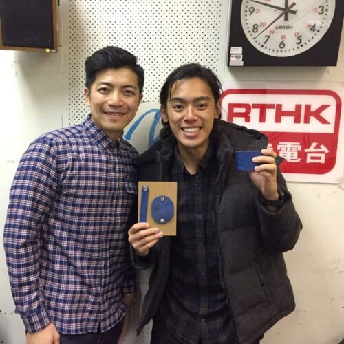 Two Asian men posing in front of a RTHK (Radio Television Hong Kong) sign, one holding a radio or audio device.
